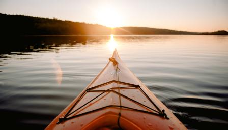 Kuvassa näkyy aurinko ja kanootin keula järvellä auringon laskiessa puiden taakse.