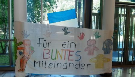 Koulun aulaan ripustettu lakana, jossa on värikkäitä kuvia ja seuraava saksankielinen teksti: Für ein buntes miteinander.