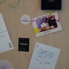 A tall bulletin board with various notes and cards attached. At the top, there is a handwritten message and a bakery advertisement featuring a dessert being eaten with a spoon. Among the cards, there are also small stickers and notes with messages like 'The panda cafe is insanely good' and 'Good luck with your new beginning!