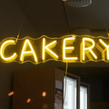 A yellow neon sign that reads 'CAKERY' hangs from the ceiling in a dimly lit space. The glow of the sign illuminates details of the room, such as a fridge and shelves in the background.